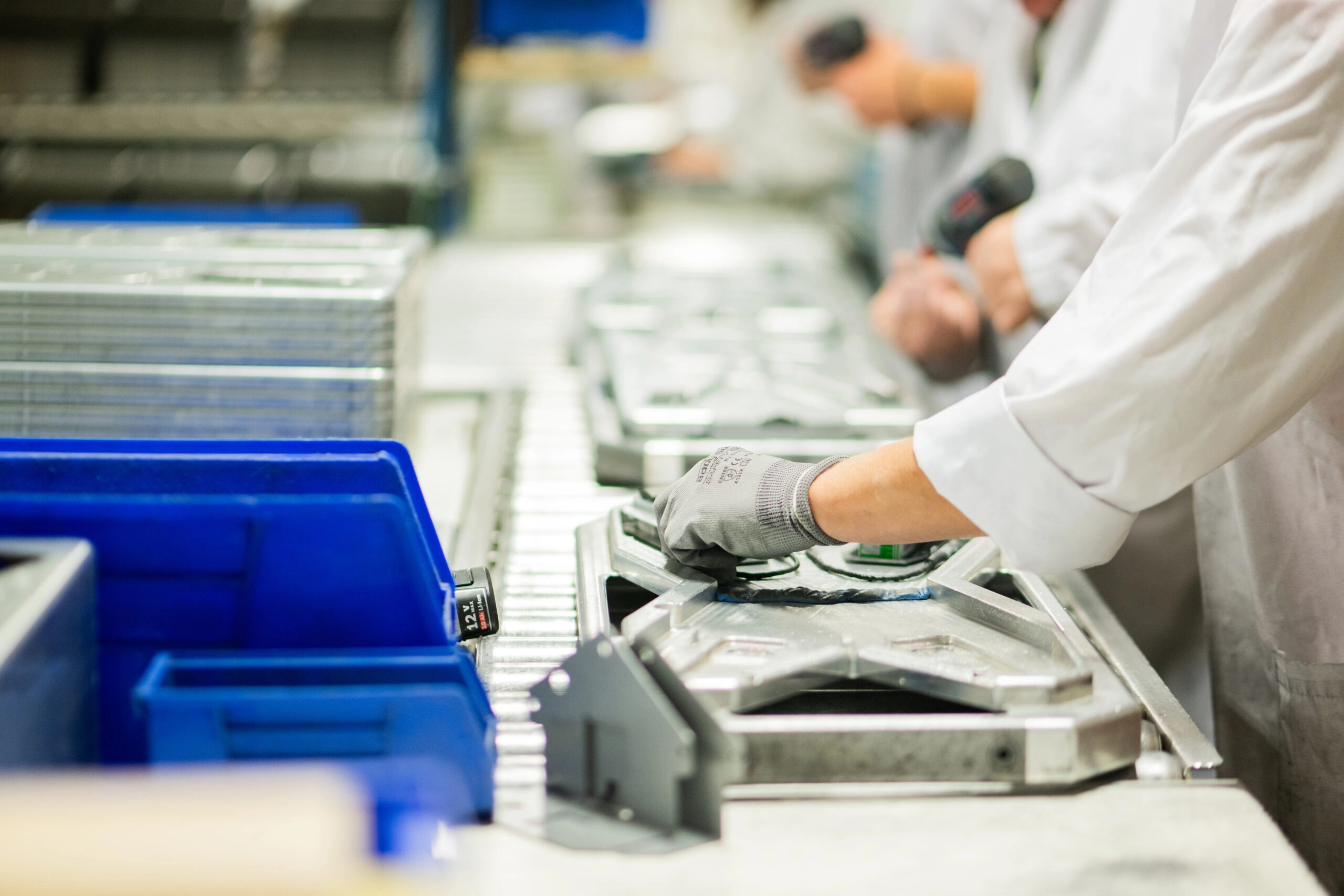 Factory worker in a white coat and gloves handling metal trays on a stainless steel production line, with blue bins nearby.