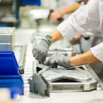 Workers in white coats and gloves assemble items on a metal production line with blue bins nearby