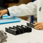 Hands of a technician in a white lab coat assembling black rectangular electronic modules on a white worktable.