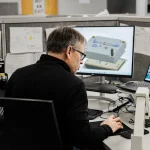 Man wearing glasses at a desk, looking at a CAD model on a large monitor in a cubicle office.