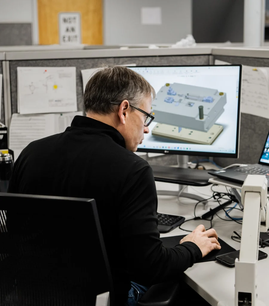 Man wearing glasses at a desk, looking at a CAD model on a large monitor in a cubicle office.